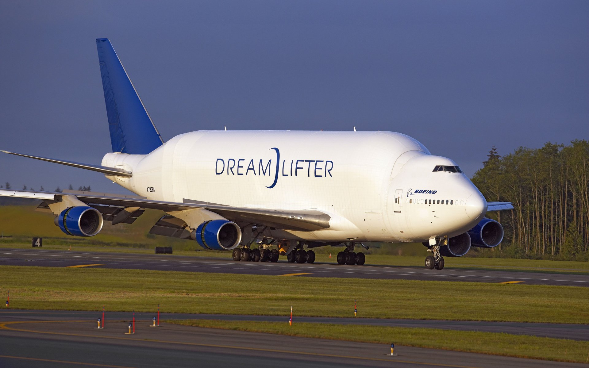 5K Ultra HD PC desktop wallpaper and background: Boeing Dreamlifter cargo aircraft taxiing on runway at dusk, white fuselage, blue tail and tree-lined horizon.