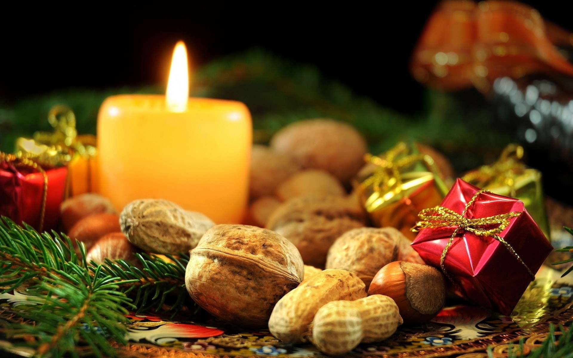A warm candle glows amidst festive nuts and small wrapped gifts, surrounded by greenery, creating a cozy holiday Christmas atmosphere in this HD desktop wallpaper.