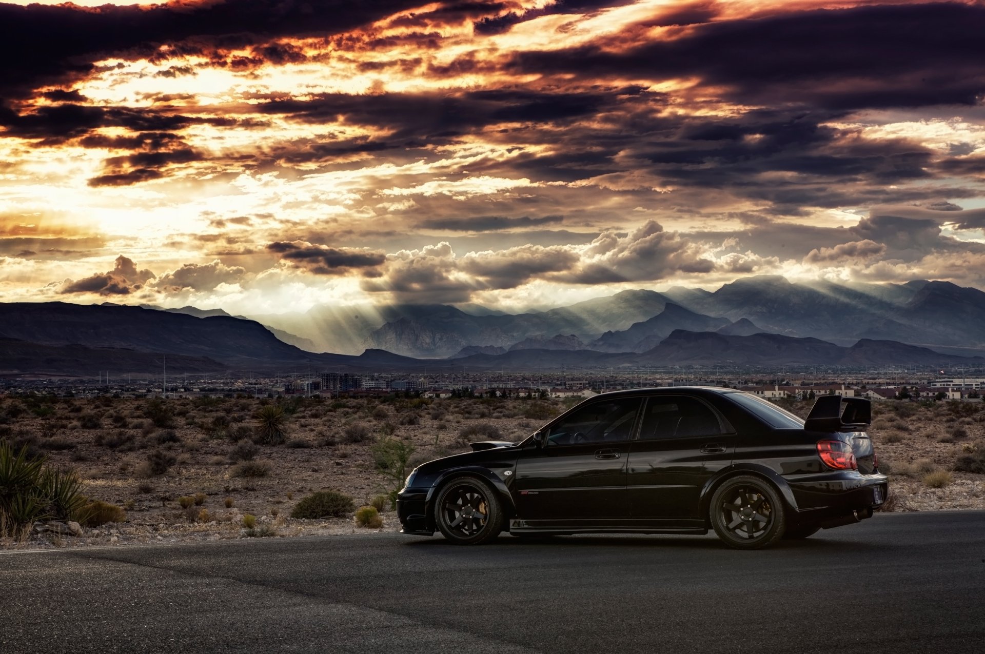 A black Subaru vehicle parked on a desert road under a dramatic, colorful sunset sky, captured in HD for a striking desktop wallpaper background.