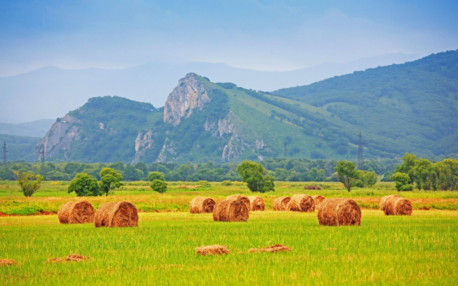 HD PC desktop wallpaper featuring a vibrant landscape with green fields, scattered hay bales, and distant rolling hills under a clear blue sky.