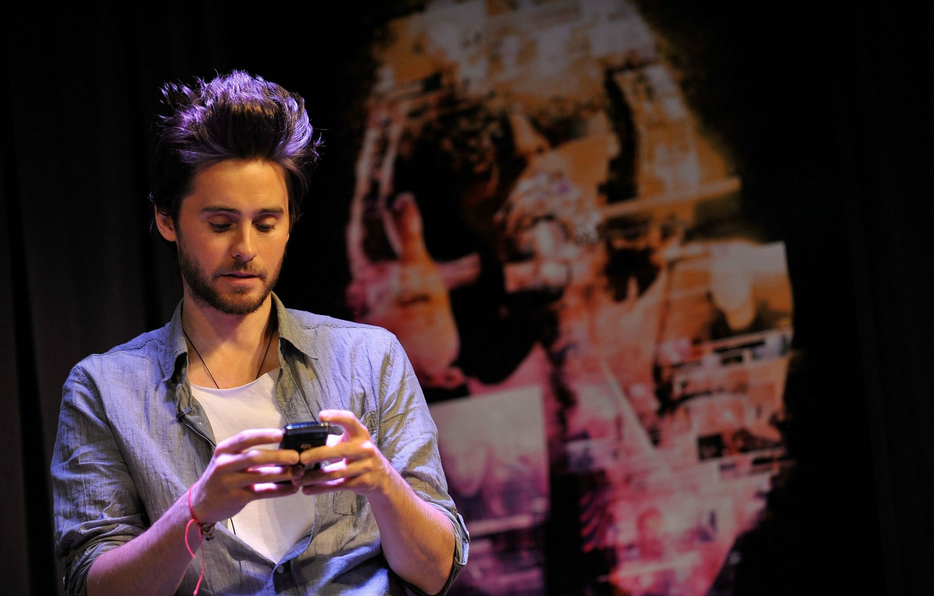 Celebrity portrait of a seated man checking his phone against a blurred poster backdrop — 2K Quad HD PC desktop wallpaper/background.