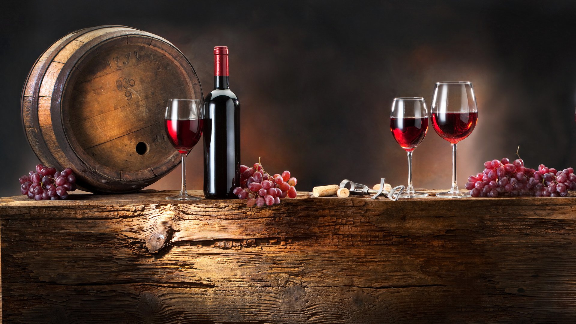 HD PC desktop wallpaper featuring a rustic wooden table with a wine barrel, a bottle of red wine, three filled glasses, and bunches of grapes against a dark background.