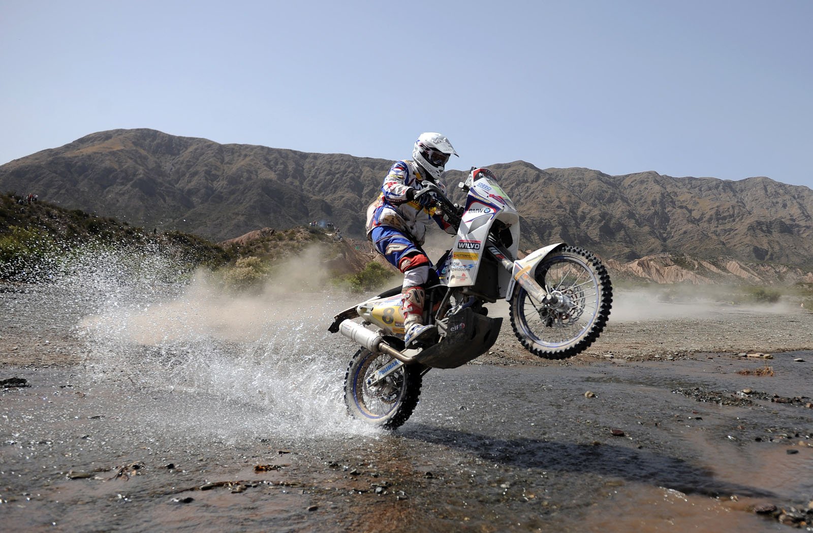 HD desktop wallpaper featuring a motorcycle rider performing a wheelie while crossing a shallow stream with mountains in the background.
