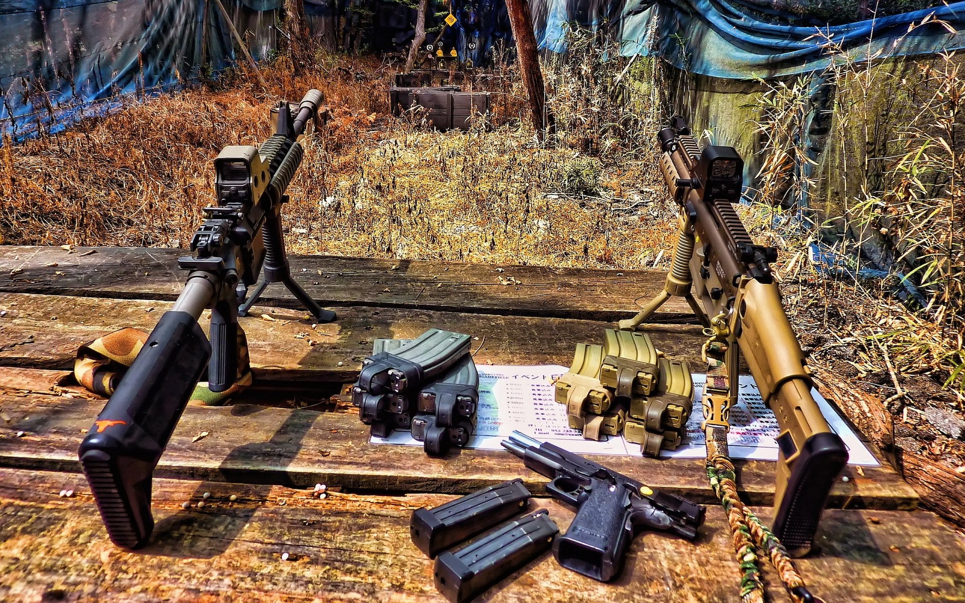 HD desktop wallpaper featuring man-made assault rifles and ammunition displayed on a wooden surface outdoors with a natural, rugged background.