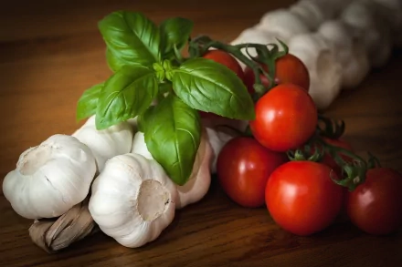 HD desktop wallpaper featuring fresh vegetables including garlic bulbs, ripe red tomatoes on the vine, and vibrant green basil leaves, displayed on a wooden surface.