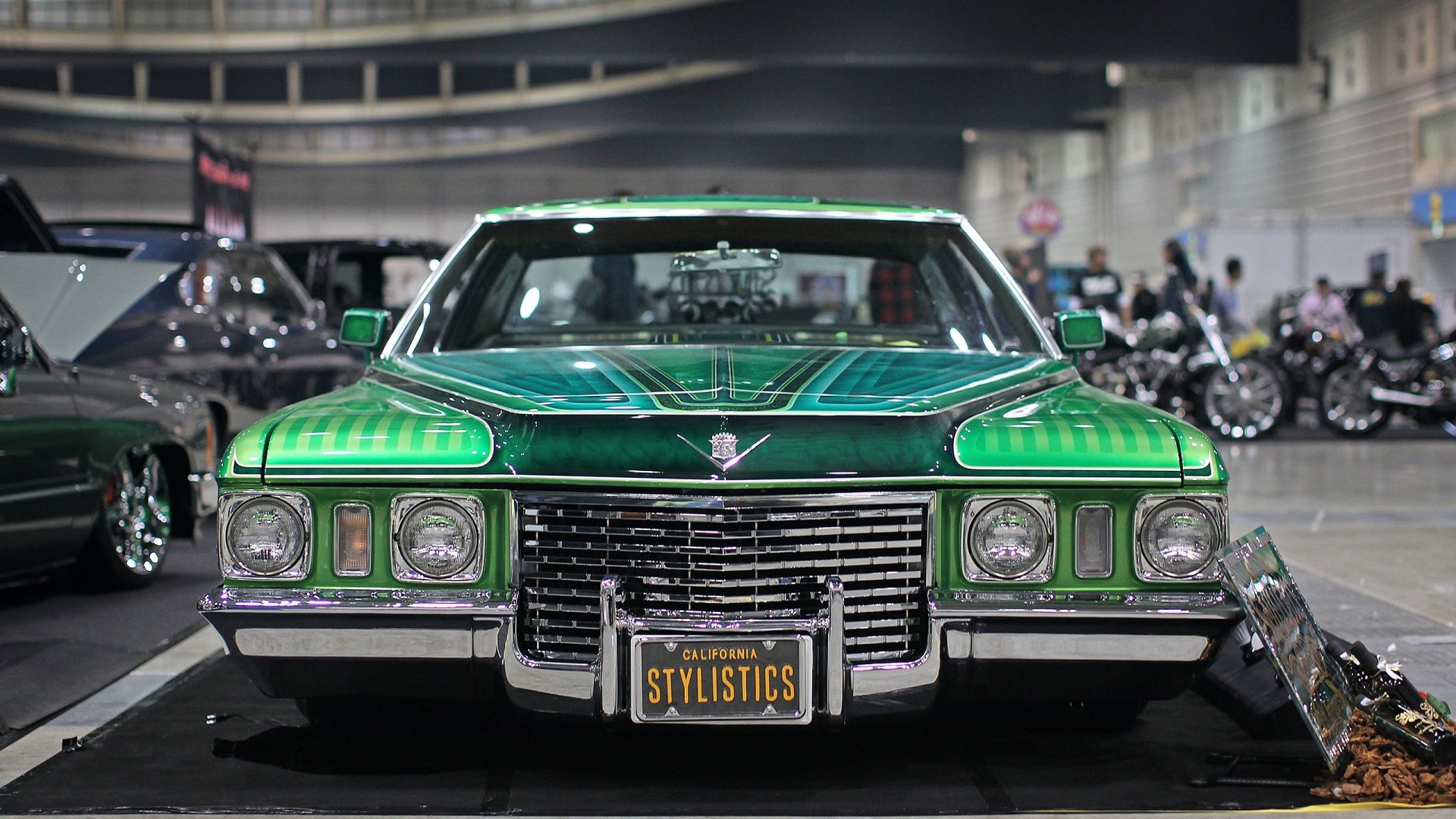 HD PC desktop wallpaper of a green Cadillac vehicle showcased indoors with a reflective floor and surrounding cars in the background.
