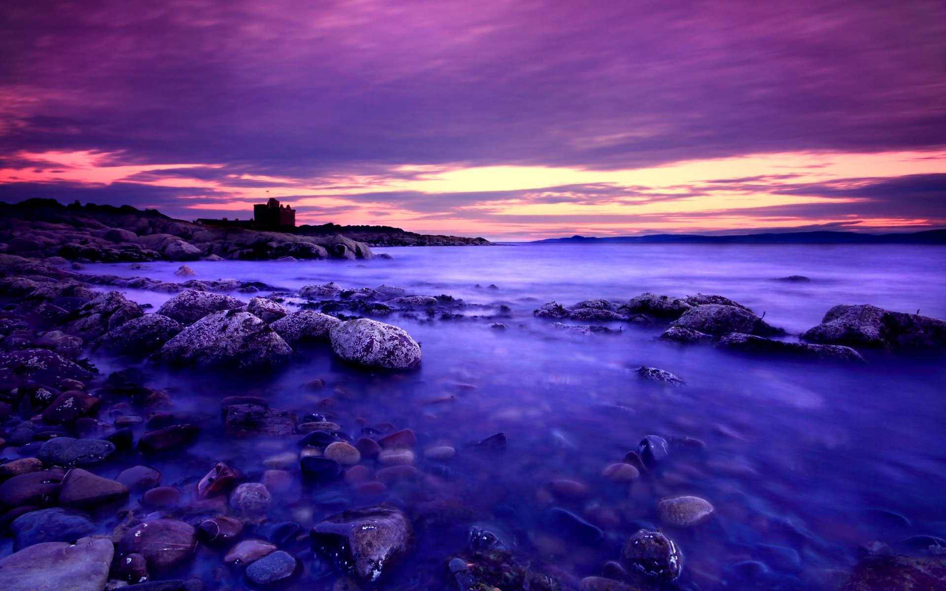 HD PC desktop wallpaper - nature coastline at dusk: rocky shore with purple-pink sky, smooth misty water and distant silhouetted building on the horizon.