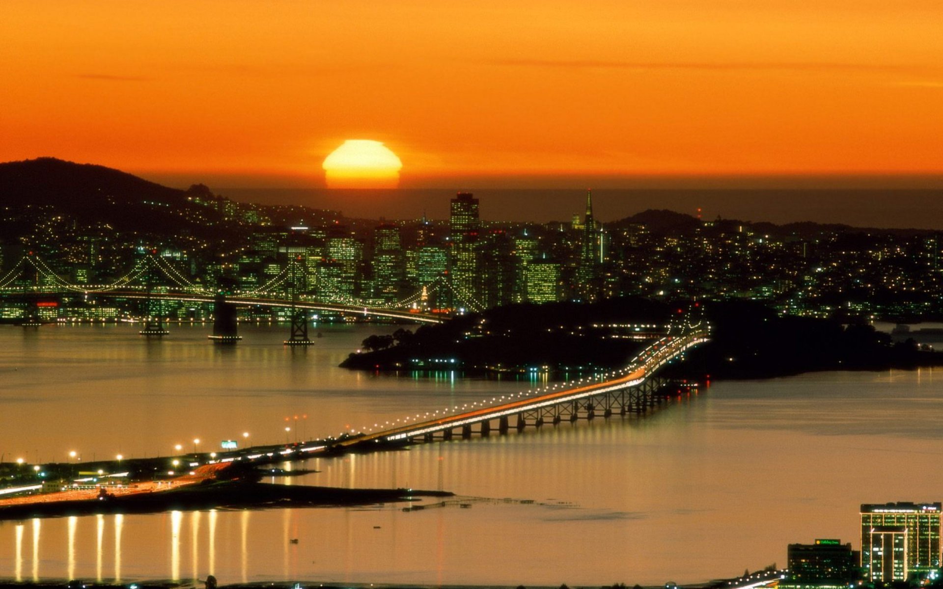 2K Quad HD PC wallpaper: San Francisco skyline with man-made Bay Bridge aglow at sunset, city lights reflecting across the calm bay.