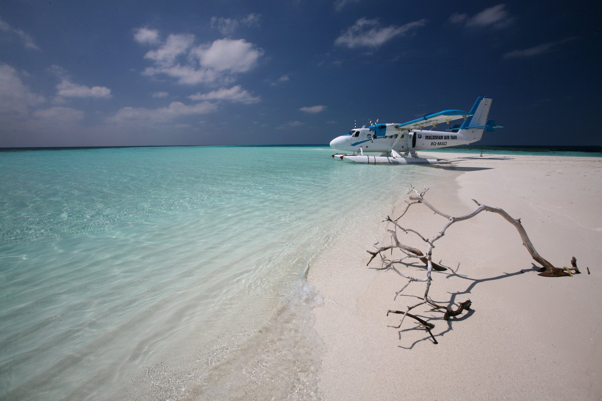 A De Havilland Canada DHC-6 Twin Otter seaplane rests on a pristine, sandy beach beside clear turquoise waters, with a stunning ocean horizon and dramatic sky in the Maldives.
