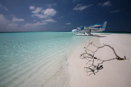A De Havilland Canada DHC-6 Twin Otter seaplane rests on a pristine, sandy beach beside clear turquoise waters, with a stunning ocean horizon and dramatic sky in the Maldives.
