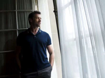 Hugh Jackman stands thoughtfully by a window, dressed in a navy polo shirt, with soft light filtering through sheer curtains. This captivating image serves as an HD desktop wallpaper.