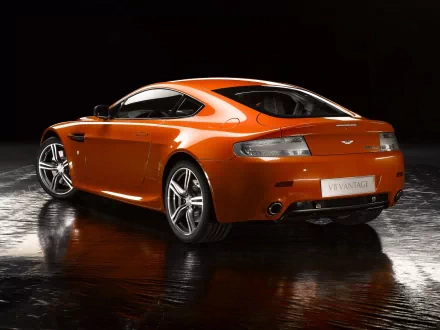 Orange Aston Martin Vantage sports car showcased as a sleek HD desktop wallpaper with a reflective floor and black background.