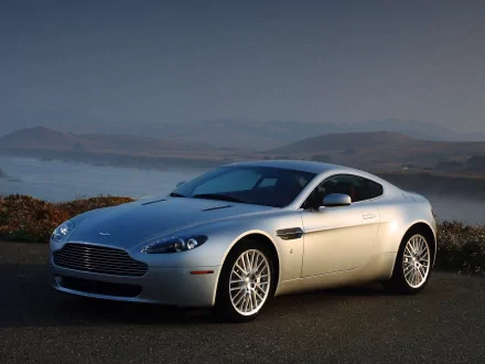 Silver Aston Martin Vantage vehicle parked on a coastal road at dusk, HD PC desktop wallpaper background with calm water and rolling hills.