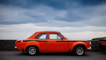 Orange classic Ford Escort side profile parked on a coastal road, 2K Quad HD PC desktop wallpaper/background.