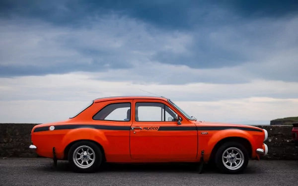 Orange classic Ford Escort side profile parked on a coastal road, 2K Quad HD PC desktop wallpaper/background.