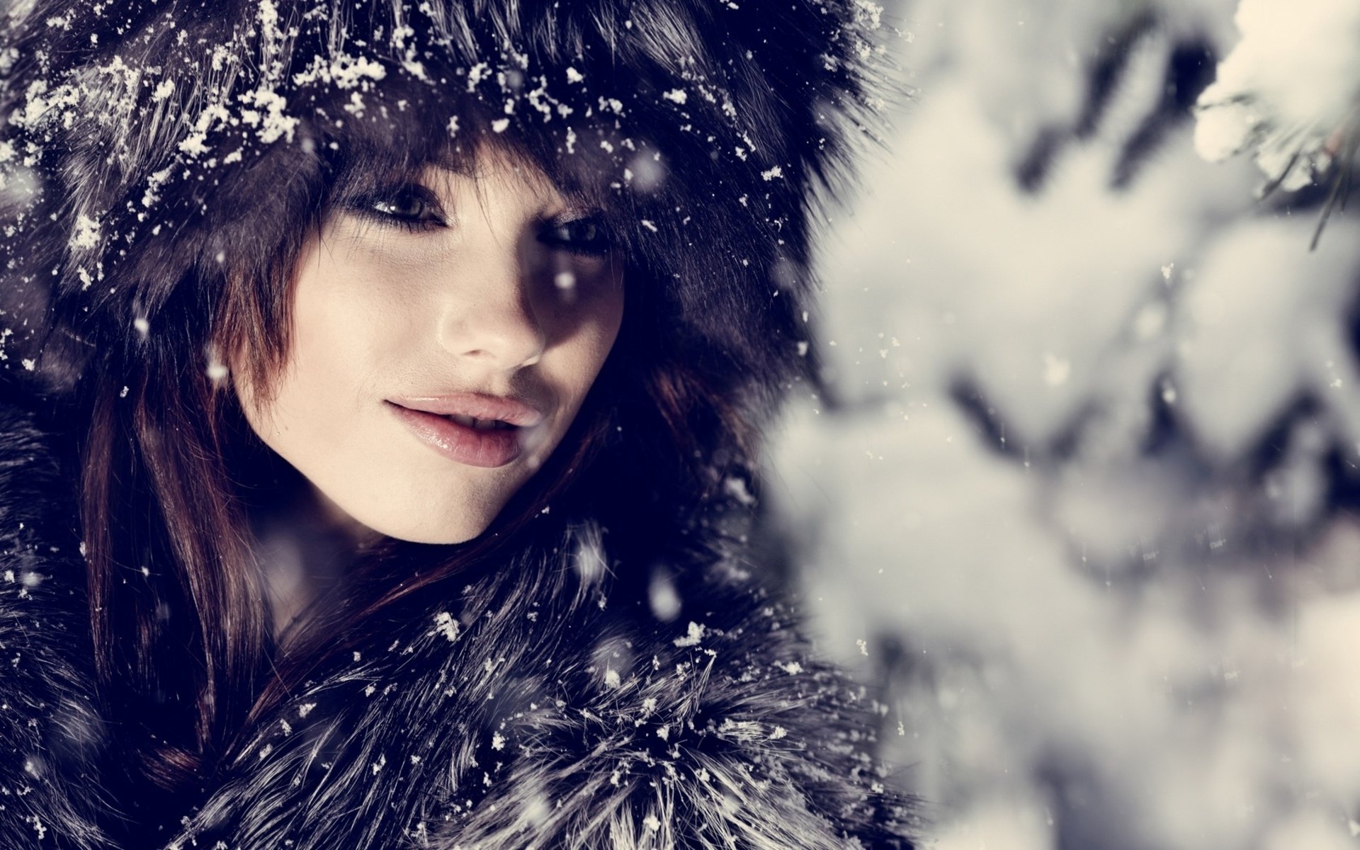 A close-up of a woman’s face, adorned in a fur-lined hat, surrounded by falling snowflakes, creating a serene winter backdrop. High-definition desktop wallpaper.