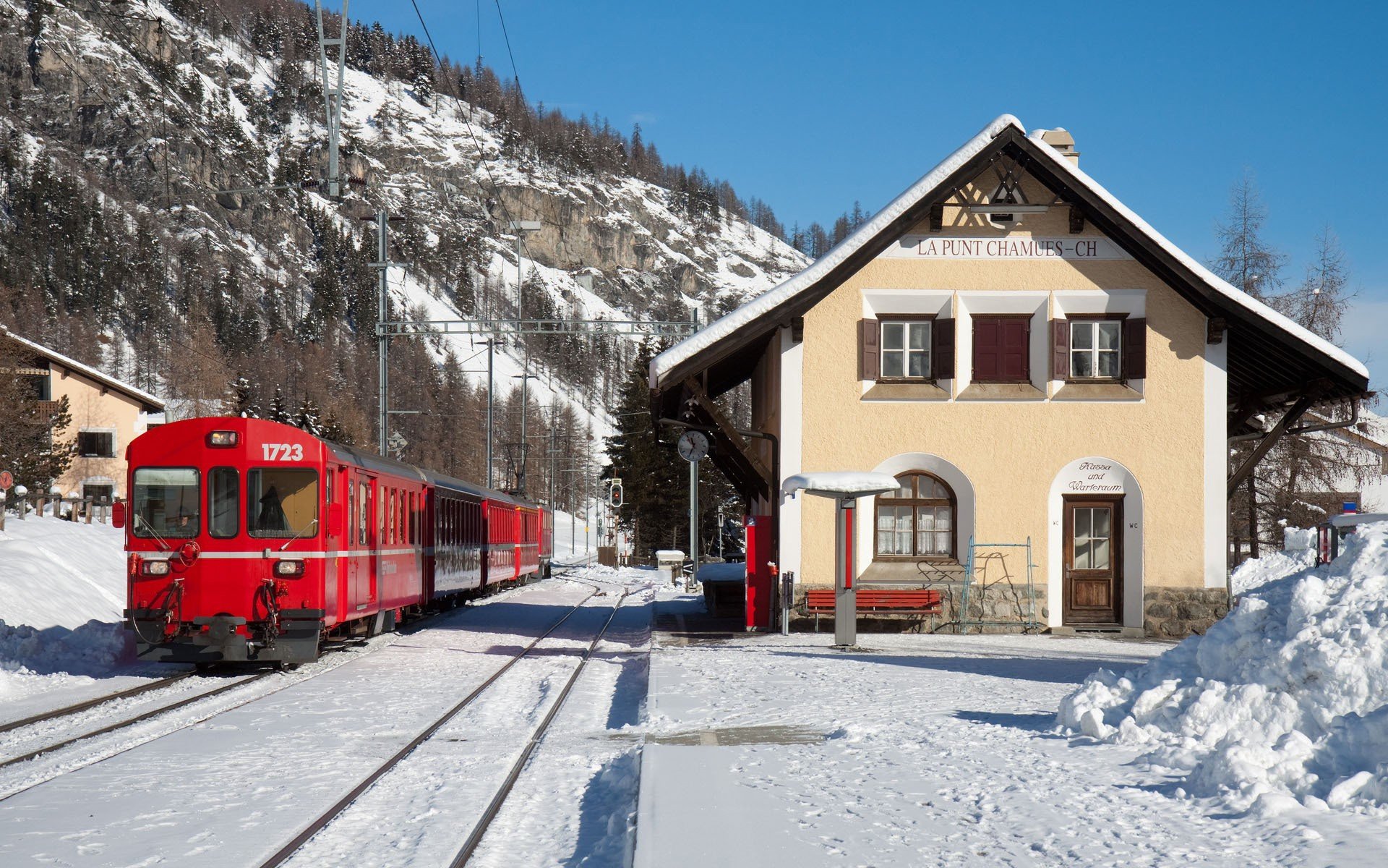 RhB train in Switzerland stopped at a snowy mountain station, captured in a high-definition desktop wallpaper showcasing a bright winter day.