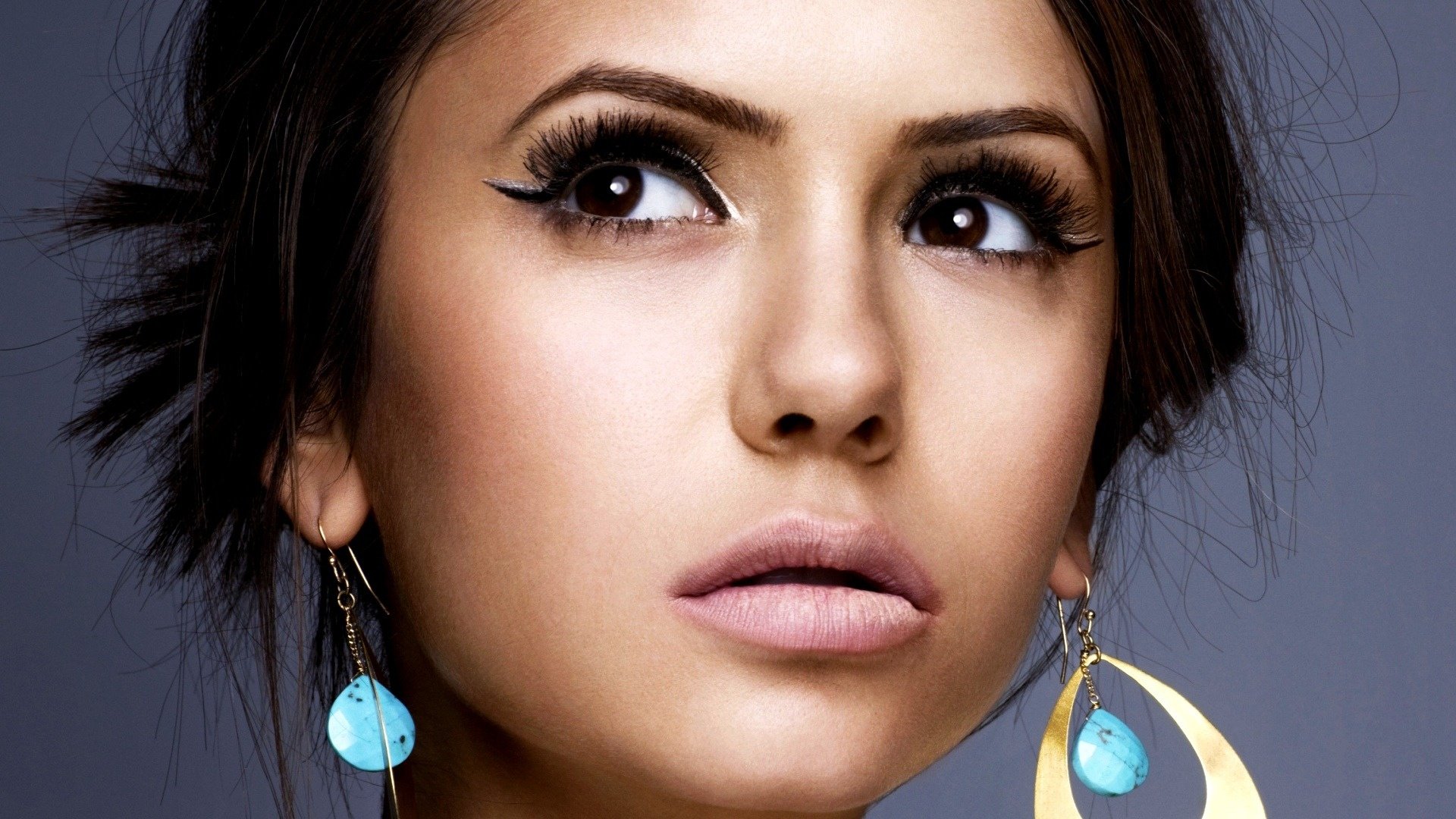 HD PC desktop wallpaper featuring a close-up of celebrity Nina Dobrev with dramatic eye makeup and distinctive blue and gold earrings.