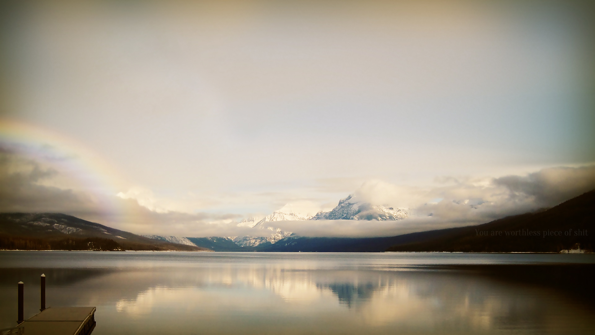 HD photography of a serene lake with mountains reflected in the calm water, captured as a desktop wallpaper featuring soft clouds and a subtle rainbow.