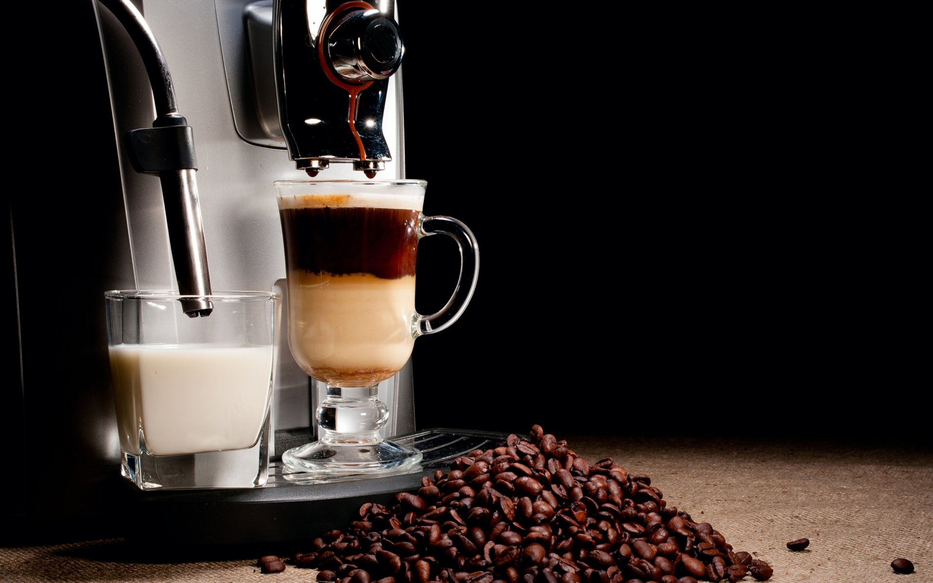 HD PC desktop wallpaper featuring a close-up of a coffee machine pouring layered coffee with milk, alongside a glass of milk and scattered coffee beans on a dark surface.