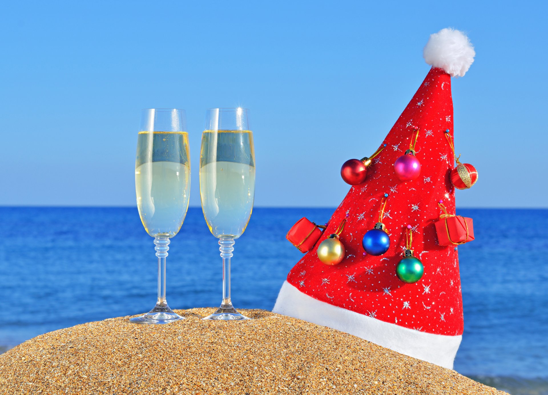 4K Ultra HD holiday-themed New Year wallpaper showing two champagne glasses and a festive red Santa hat adorned with ornaments on a sandy beach with a blue ocean backdrop.
