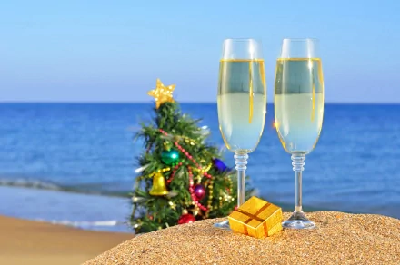 4K Ultra HD desktop wallpaper featuring two champagne glasses, a small decorated Christmas tree, and a gift box on a sandy beach with the ocean in the background for New Year holiday.