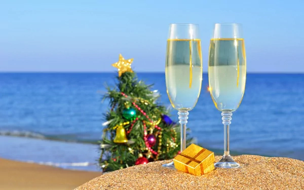 4K Ultra HD desktop wallpaper featuring two champagne glasses, a small decorated Christmas tree, and a gift box on a sandy beach with the ocean in the background for New Year holiday.
