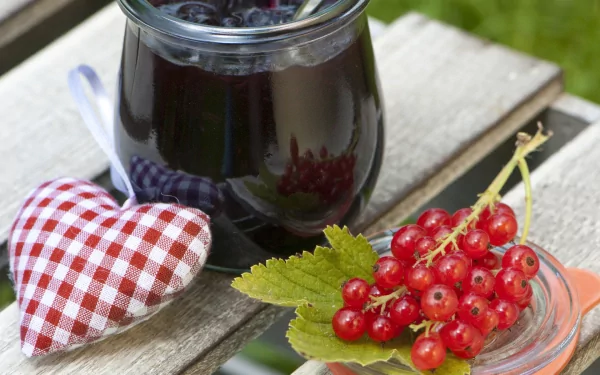 HD PC desktop wallpaper: close-up of a jar of dark jam, fresh red currants on a leaf, and a red-checked fabric heart resting on a weathered wooden bench.