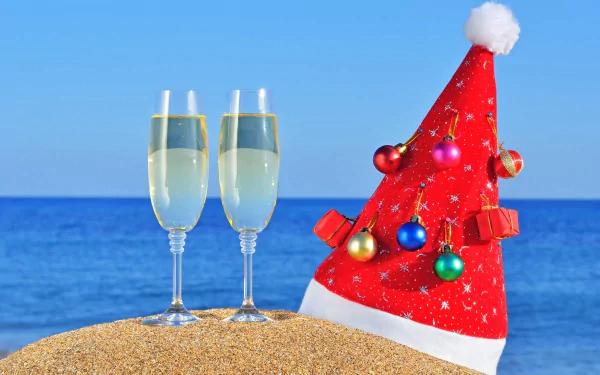 4K Ultra HD holiday-themed New Year wallpaper showing two champagne glasses and a festive red Santa hat adorned with ornaments on a sandy beach with a blue ocean backdrop.