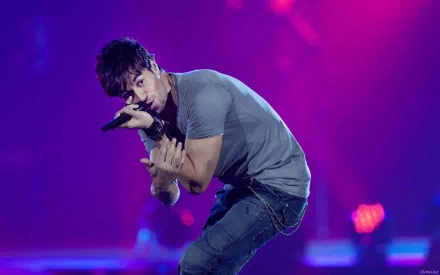 HD desktop wallpaper of Enrique Iglesias performing on stage, holding a microphone and leaning forward with a vibrant purple and blue lit background.