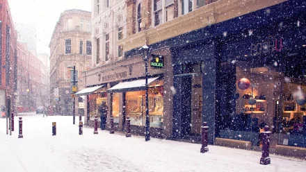 HD desktop wallpaper of a charming winter scene, capturing snow falling gently on a city street lined with shops and old buildings.