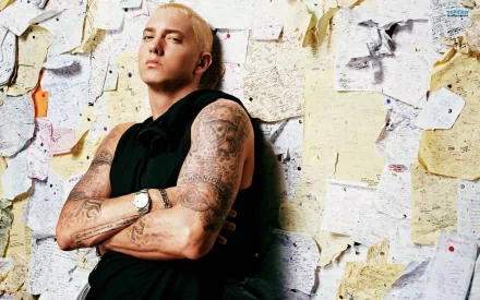 Eminem poses confidently against a backdrop of notes and lyrics, showcasing his signature tattoos. This HD wallpaper captures the essence of his music and artistry.