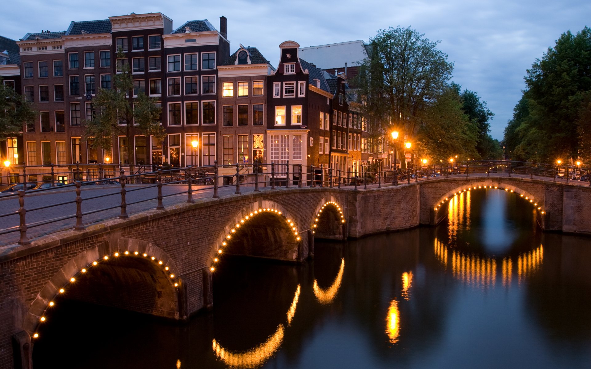 Amsterdam, Netherlands canal at dusk with a man-made arched bridge lit by string lights reflecting in water — 2K Quad HD PC desktop wallpaper/background