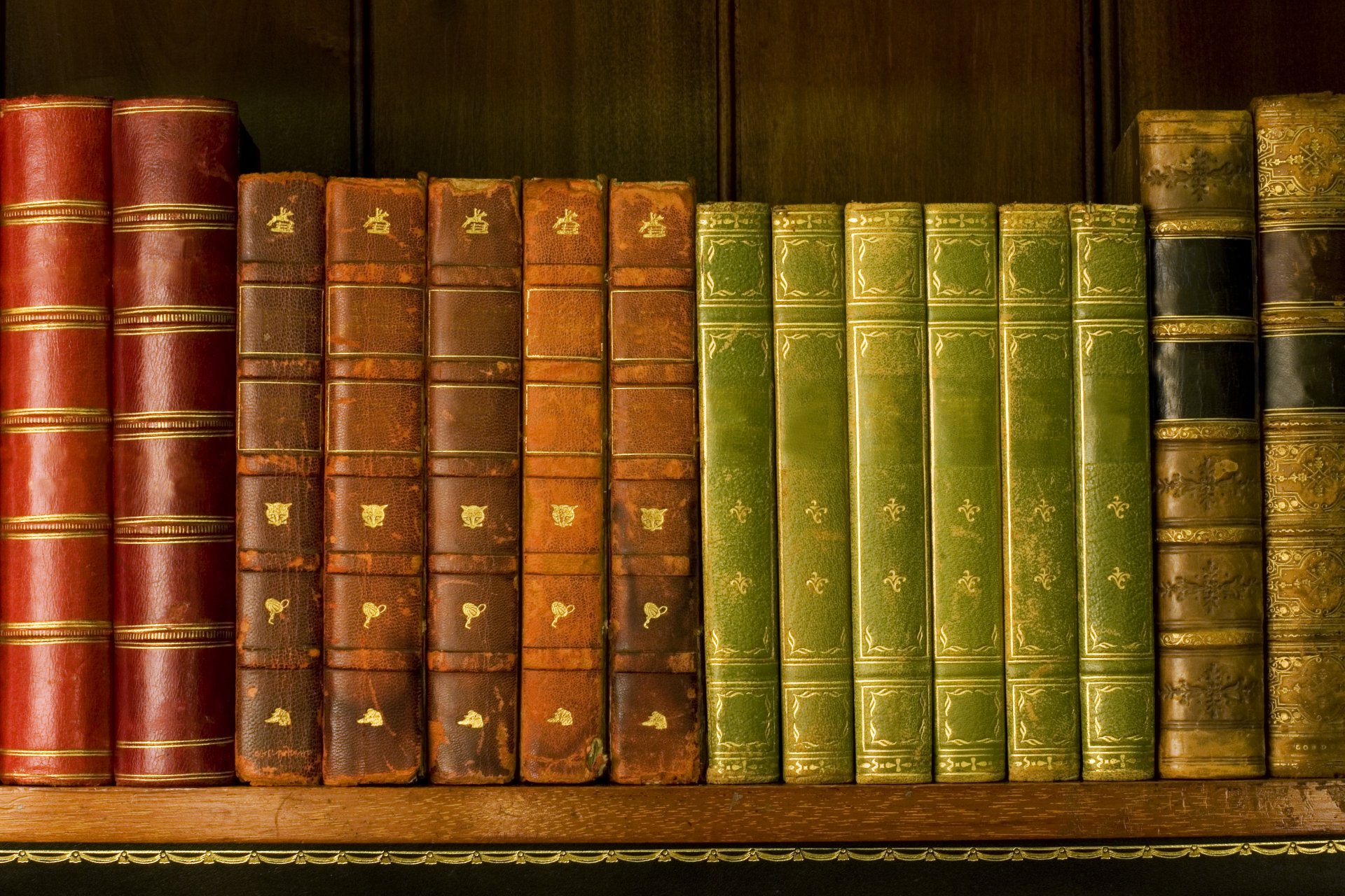 A collection of antique books displayed on a wooden shelf, showcasing rich hues of red, brown, and green, creating a warm and inviting atmosphere for a desktop wallpaper.