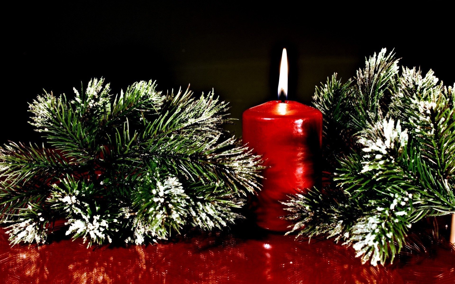 A warm red candle surrounded by snowy evergreen branches, creating a festive atmosphere for the holiday season in this HD Christmas-themed wallpaper.