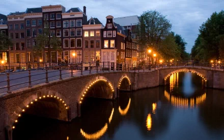 Amsterdam, Netherlands canal at dusk with a man-made arched bridge lit by string lights reflecting in water — 2K Quad HD PC desktop wallpaper/background