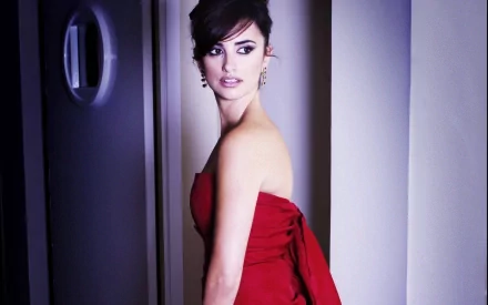 A striking HD desktop wallpaper featuring Penelope Cruz in a red strapless dress, posing elegantly as she looks over her shoulder against a soft-lit backdrop.