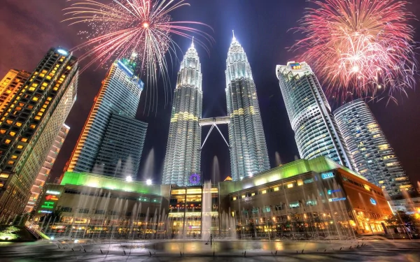 Fireworks light up the night sky over Kuala Lumpur's iconic Petronas Towers and surrounding skyscrapers, with a fountain and city buildings in view.