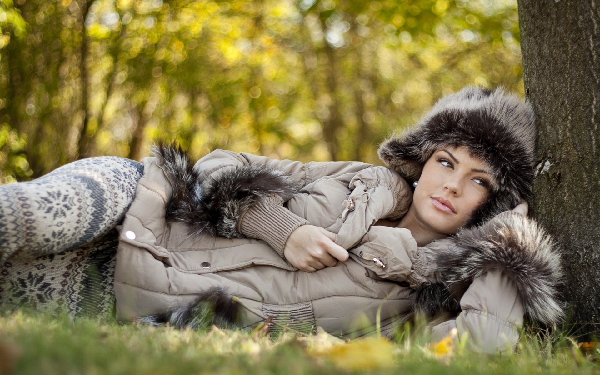 A beautiful woman relaxes against a tree in autumn, wearing a cozy fur-trimmed jacket and patterned leggings. The vibrant greenery and soft light create a serene atmosphere.