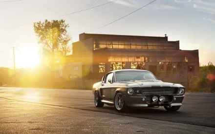HD desktop wallpaper featuring a classic Ford Mustang parked on a deserted road, with the sun setting behind an industrial building in the background.