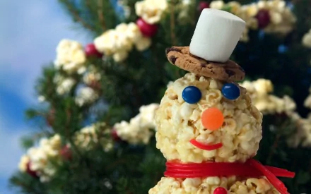 HD PC desktop wallpaper of a popcorn snowman wearing a cookie hat topped with a marshmallow, candy eyes and nose, red ribbon scarf, with a festive Christmas tree blurred in the background.