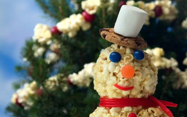 HD PC desktop wallpaper of a popcorn snowman wearing a cookie hat topped with a marshmallow, candy eyes and nose, red ribbon scarf, with a festive Christmas tree blurred in the background.