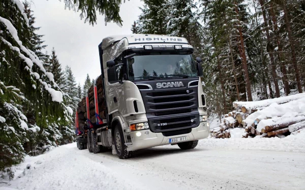 HD desktop wallpaper featuring a Scania truck driving on a snowy forest road surrounded by tall pine trees and stacked logs.