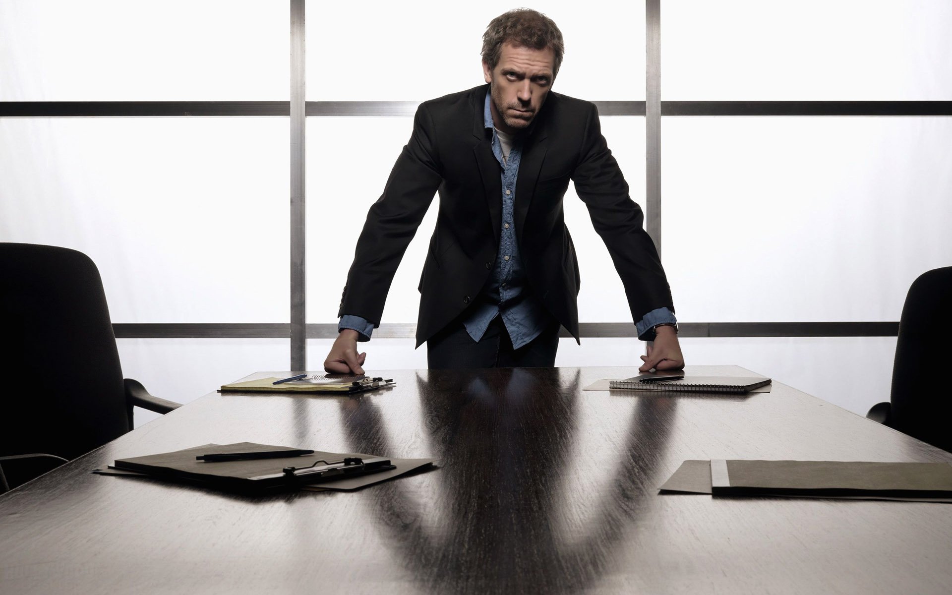 HD desktop wallpaper of a celebrity actor in a suit leaning over a conference table, backlit window panels behind him and folders spread on the table.