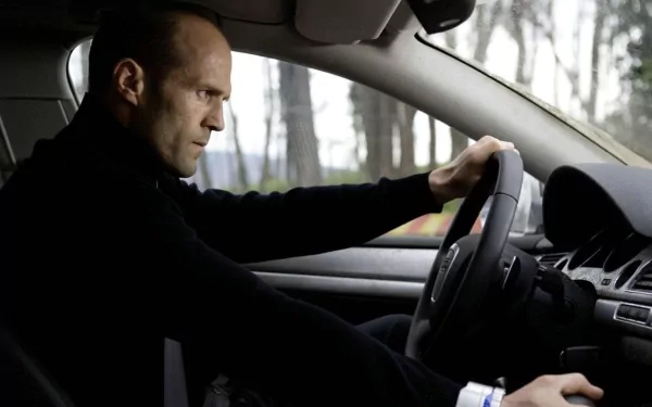 HD wallpaper of a focused driver at the wheel from The Transporter, set against a wooded backdrop.