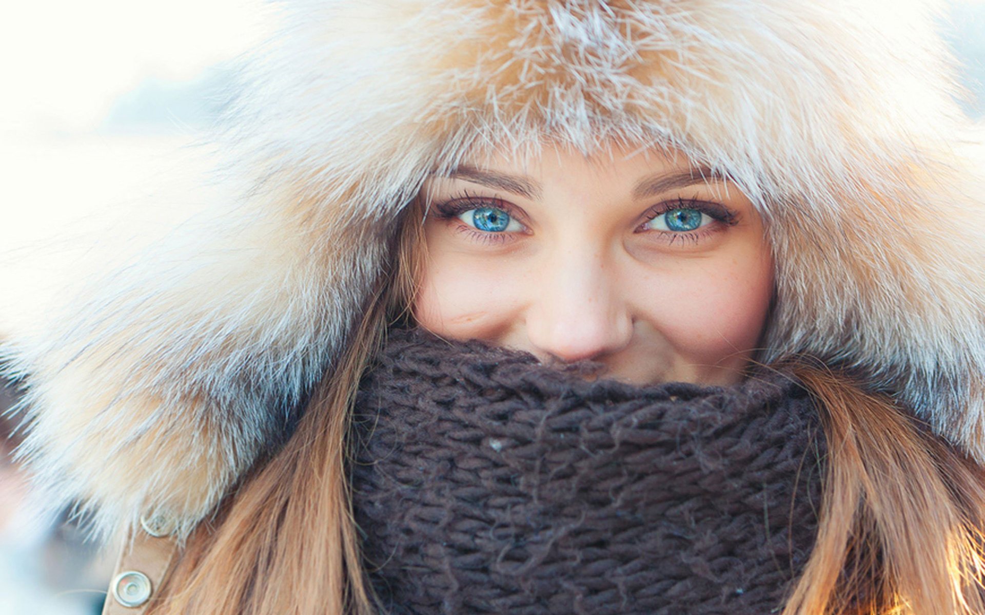 HD wallpaper of a woman with bright blue eyes, her face framed by a fur-lined hood and a dark knitted scarf, creating a cozy winter look.