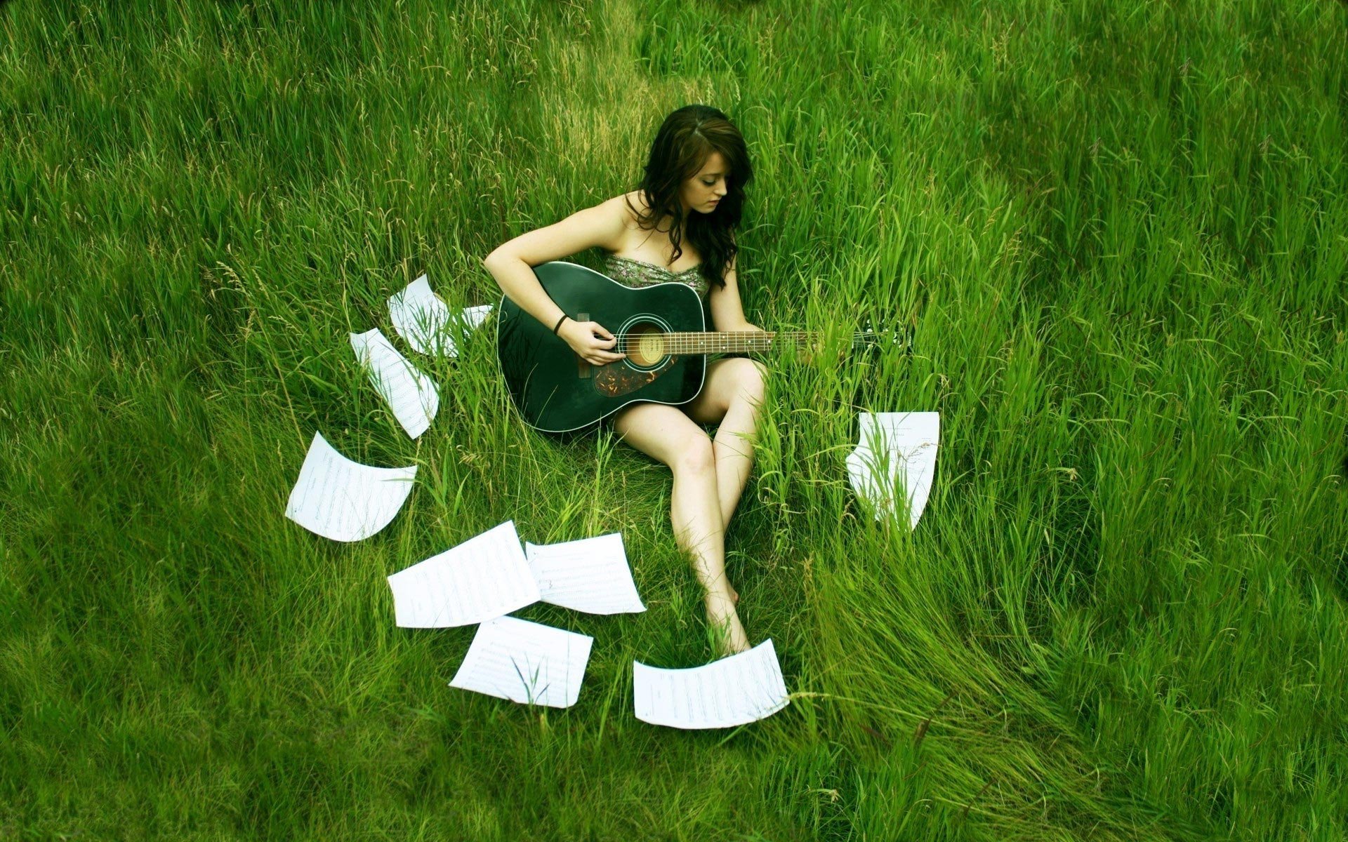 A woman with long hair sits in green grass, playing a guitar surrounded by scattered music sheets, capturing a serene and artistic mood in this HD desktop wallpaper.