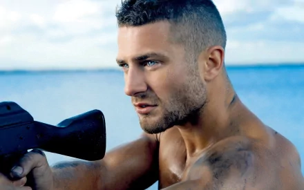 A handsome man with a strong build and intense expression holds a weapon, set against a backdrop of tranquil water and sky. This HD image makes a striking desktop wallpaper.