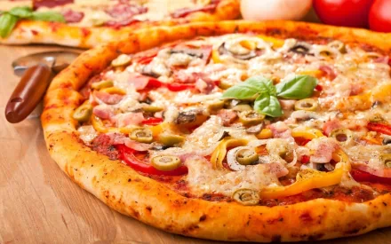 HD PC desktop wallpaper of a freshly baked pizza topped with olives, bell peppers, mushrooms, and basil on a wooden surface with tomatoes in the background.