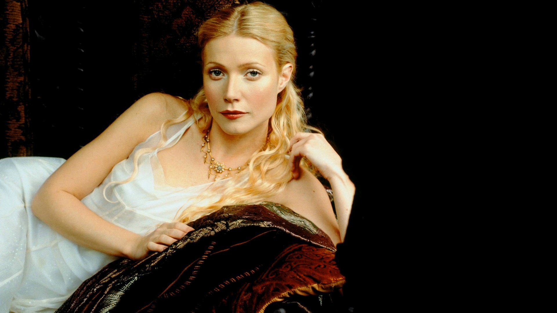 HD PC desktop wallpaper featuring celebrity Gwyneth Paltrow reclining on a dark, ornate sofa, dressed in a white, delicate top with soft lighting highlighting her face.